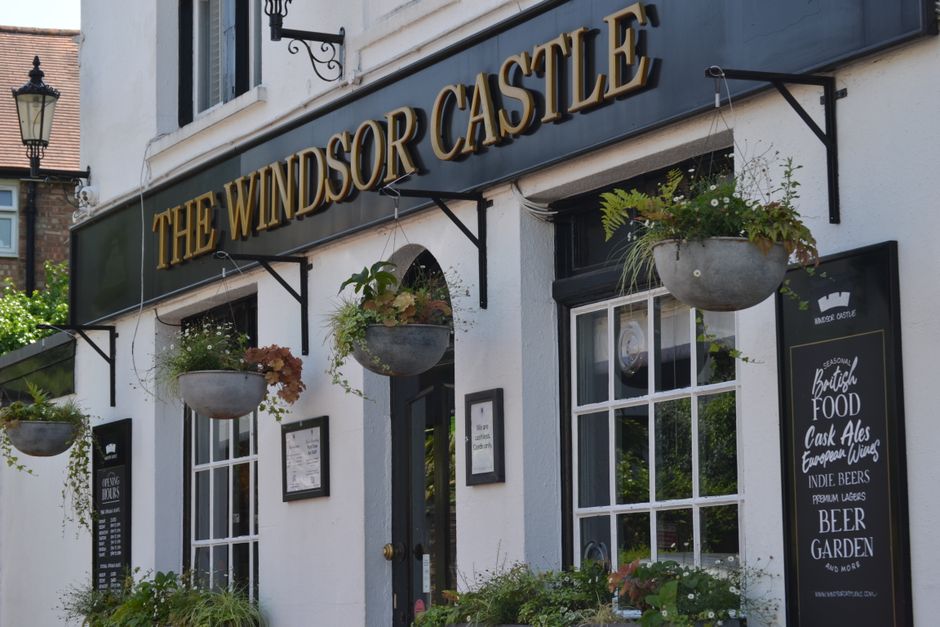 A floral arrangement in a round, gray ceramic pot is hanging outside the white exterior of The Windsor Castle pub, associated with Hertford Castle. The arrangement includes a mix of greenery, ferns, a
