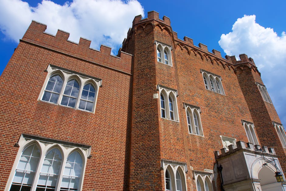 A close-up view of Hertford Castle's brick facade, featuring tall, narrow, white-framed windows with pointed arches, set against a bright blue sky with scattered white clouds. The castle's red-brick e